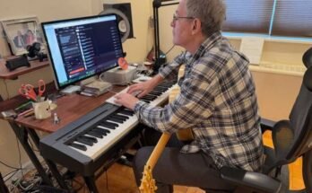 En esta foto, John Beltzer de Songs of Love Foundation está en un teclado de piano electrónico y golpea un teclado de computadora que está arriba. Mira a un instructor de computadora que está en un escritorio detrás del teclado de piano. Una guitarra descansa en su regazo.