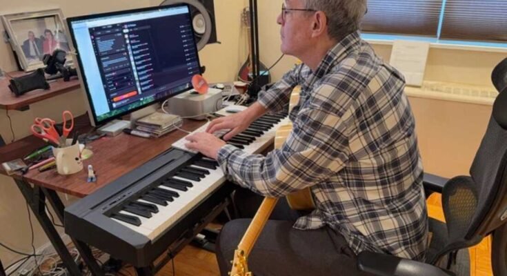 En esta foto, John Beltzer de Songs of Love Foundation está en un teclado de piano electrónico y golpea un teclado de computadora que está arriba. Mira a un instructor de computadora que está en un escritorio detrás del teclado de piano. Una guitarra descansa en su regazo.