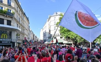 Una bandera blanca con una gran rebanada de sandía está agitado durante la línea roja para el evento Gaza en París el 8 de julio. La mayoría de los manifestantes usan camisas rojas, y caminan en una calle llena de edificios con varios estados.