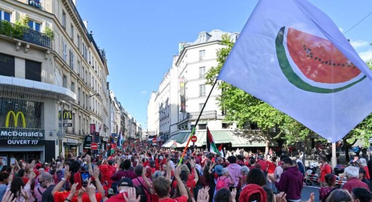 Una bandera blanca con una gran rebanada de sandía está agitado durante la línea roja para el evento Gaza en París el 8 de julio. La mayoría de los manifestantes usan camisas rojas, y caminan en una calle llena de edificios con varios estados.